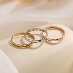 May include: Three thin metal rings in gold, silver, and rose gold tones. The gold ring is engraved with the text forever yours, the silver ring features the text milo and 10.21.23, and the rose gold ring has a smooth, plain polished surface.