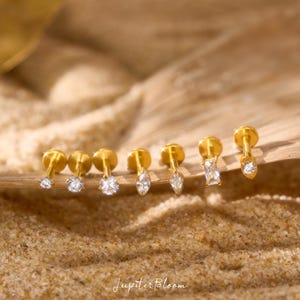 May include: A selection of gold stud earrings featuring clear gemstones in different shapes. The earrings are arranged on a light brown surface with a sandy texture. The brand name "Jupiter Bloom" is visible at the bottom.