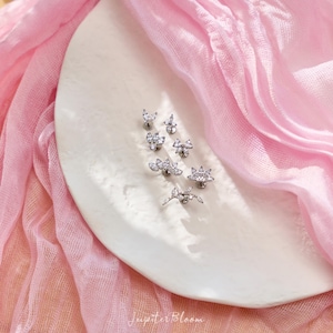 May include: An assortment of silver stud earrings, each adorned with clear gemstones in floral and geometric patterns. The earrings are presented on a white, oval plate, set against a backdrop of soft pink fabric. The text "Jupiter Bloom" is visible.