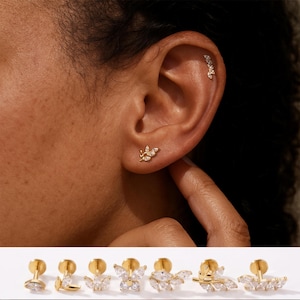 May include: Gold-toned earrings with clear gemstones. One earring features a leaf design, while another has a row of small, triangular stones. Additional earring styles are displayed below, showcasing various shapes and gemstone arrangements.