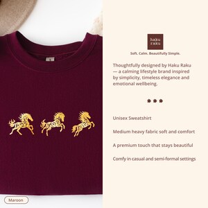 Op de afbeelding: Bordeaux unisex sweatshirt met drie gouden paardenontwerpen. De sweatshirt heeft de tekst "haku raku" en het woord "Maroon" onderaan. Het ontwerp wordt beschreven als een rustgevend lifestyle merk ge&iuml;nspireerd door eenvoud, tijdloze elegantie en emotioneel welzijn.