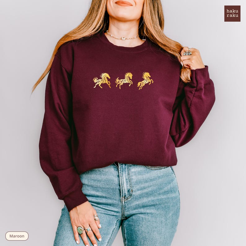 Op de afbeelding: Bordeaux sweatshirt met drie gouden geborduurde paardenontwerpen. De top met ronde hals en lange mouwen wordt gecombineerd met een blauwe jeans. Het woord "Maroon" staat linksonder in de afbeelding.