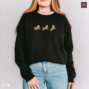 Op de afbeelding: Zwart sweatshirt met drie gouden geborduurde paardenmotieven. Het sweatshirt heeft een ronde hals en lange mouwen. De merknaam "haku raku" staat rechtsboven. Het woord "Black" staat linksonder.