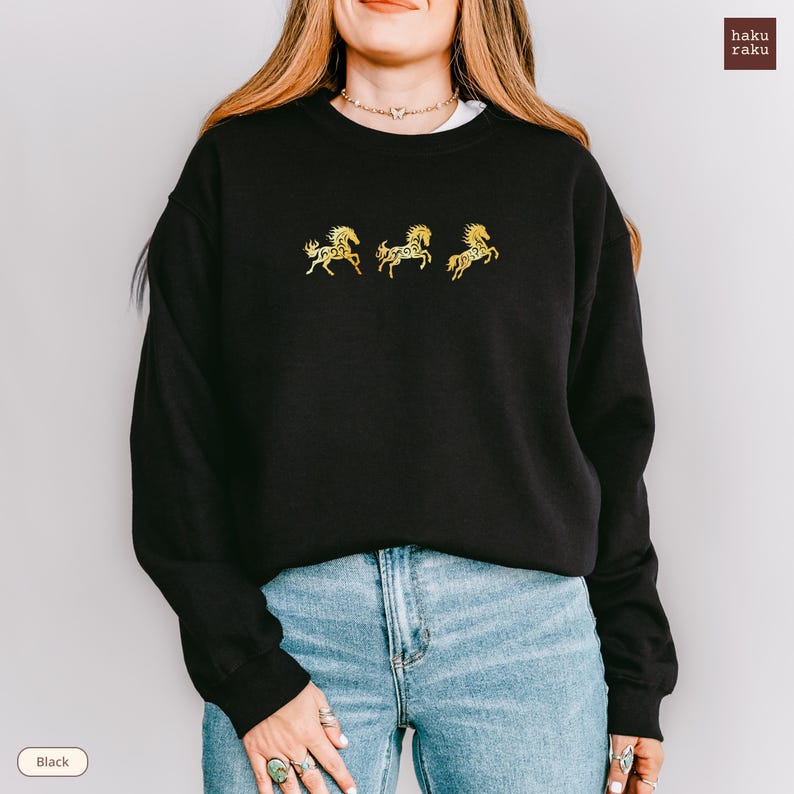 Op de afbeelding: Zwart sweatshirt met drie gouden geborduurde paardenmotieven. Het sweatshirt heeft een ronde hals en lange mouwen. De merknaam "haku raku" staat rechtsboven. Het woord "Black" staat linksonder.