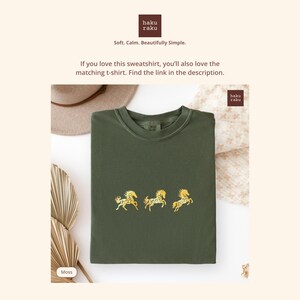 Op de afbeelding: Een mosgroene sweatshirt met drie goud geborduurde paardenontwerpen. De sweatshirt heeft een ronde hals en een relaxte pasvorm. Het merklogo staat in de rechterbovenhoek. De tekst op de afbeelding luidt "Soft. Calm. Beautifully Simple."
