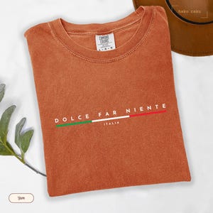 May include: A folded, burnt orange t-shirt with the words "DOLCE FAR NIENTE" and "ITALIA" printed on the front. The text is accompanied by a horizontal line in the colors of the Italian flag. The shirt is made of a soft material.