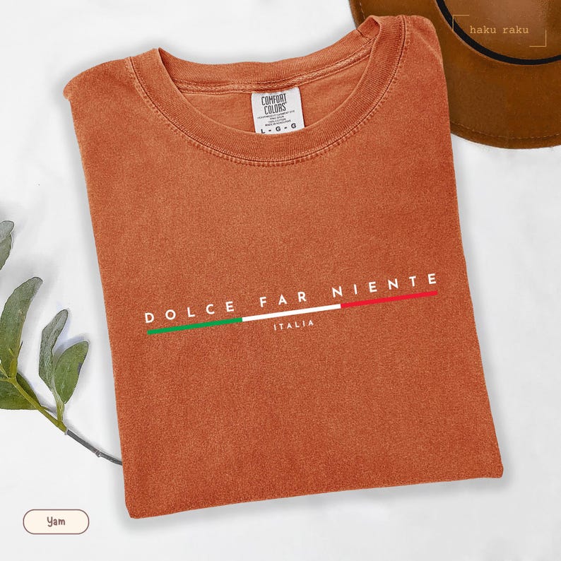 May include: A folded, burnt orange t-shirt with the words "DOLCE FAR NIENTE" and "ITALIA" printed on the front. The text is accompanied by a horizontal line in the colors of the Italian flag. The shirt is made of a soft material.