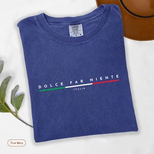 May include: A navy blue t-shirt with the words "DOLCE FAR NIENTE" and "ITALIA" printed in white. The design includes a horizontal line in the colors of the Italian flag. The shirt is folded and has a tag that says "COMFORT COLORS".