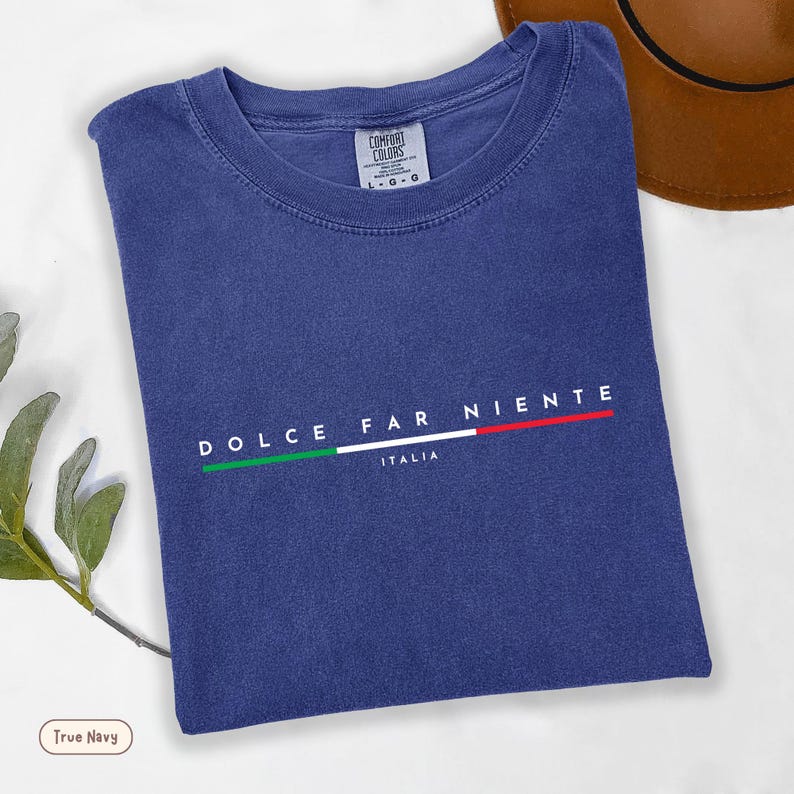 May include: A navy blue t-shirt with the words "DOLCE FAR NIENTE" and "ITALIA" printed in white. The design includes a horizontal line in the colors of the Italian flag. The shirt is folded and has a tag that says "COMFORT COLORS".