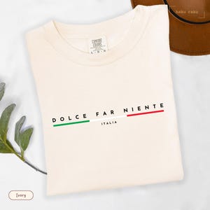 May include: Ivory-colored t-shirt with the words "DOLCE FAR NIENTE" in black, and "ITALIA" below. The design includes green, white, and red stripes, representing the Italian flag. The shirt is made of a soft material.