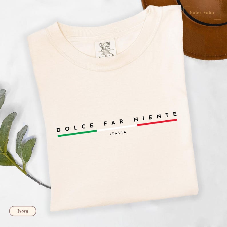 May include: Ivory-colored t-shirt with the words "DOLCE FAR NIENTE" in black, and "ITALIA" below. The design includes green, white, and red stripes, representing the Italian flag. The shirt is made of a soft material.