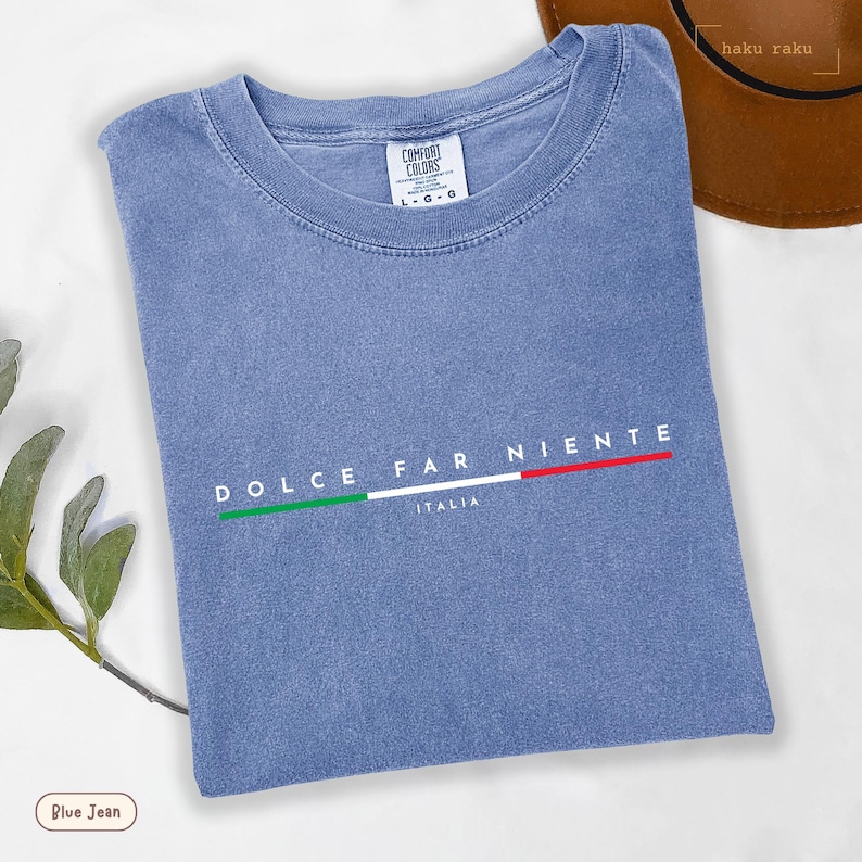 May include: A blue jean colored t-shirt with the words "DOLCE FAR NIENTE" and "ITALIA" printed on the front. The shirt has a crew neck and the Italian flag is printed above the word "ITALIA". The shirt is made by Comfort Colors.