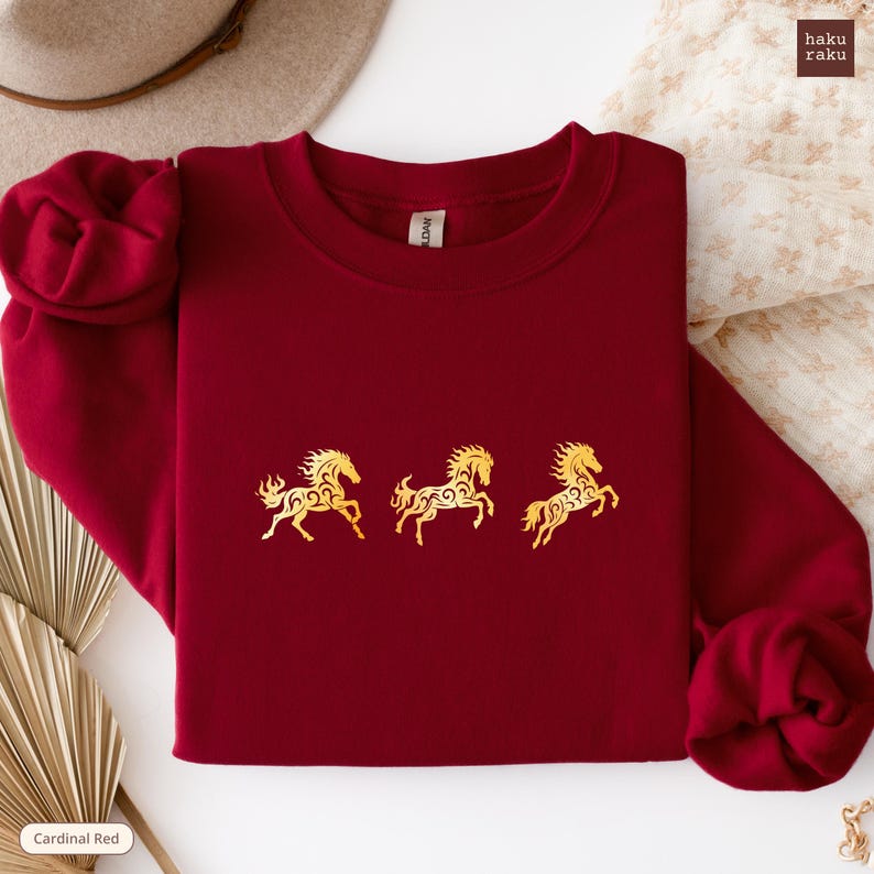 Op de afbeelding: Een bordeauxrode sweatshirt met drie gouden paardenmotieven. De paarden zijn gestileerd met ingewikkelde details. De sweatshirt heeft een ronde hals en een label met "Gildan". De woorden "haku raku" staan rechtsboven.