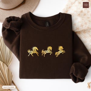 Op de afbeelding: Donkerchocoladekleurige sweatshirt met drie goudkleurige paardenontwerpen. De paarden zijn gestileerd met ingewikkelde patronen. De sweatshirt is opgevouwen, met de mouwen en halslijn zichtbaar. De merknaam "haku raku" staat rechtsboven.