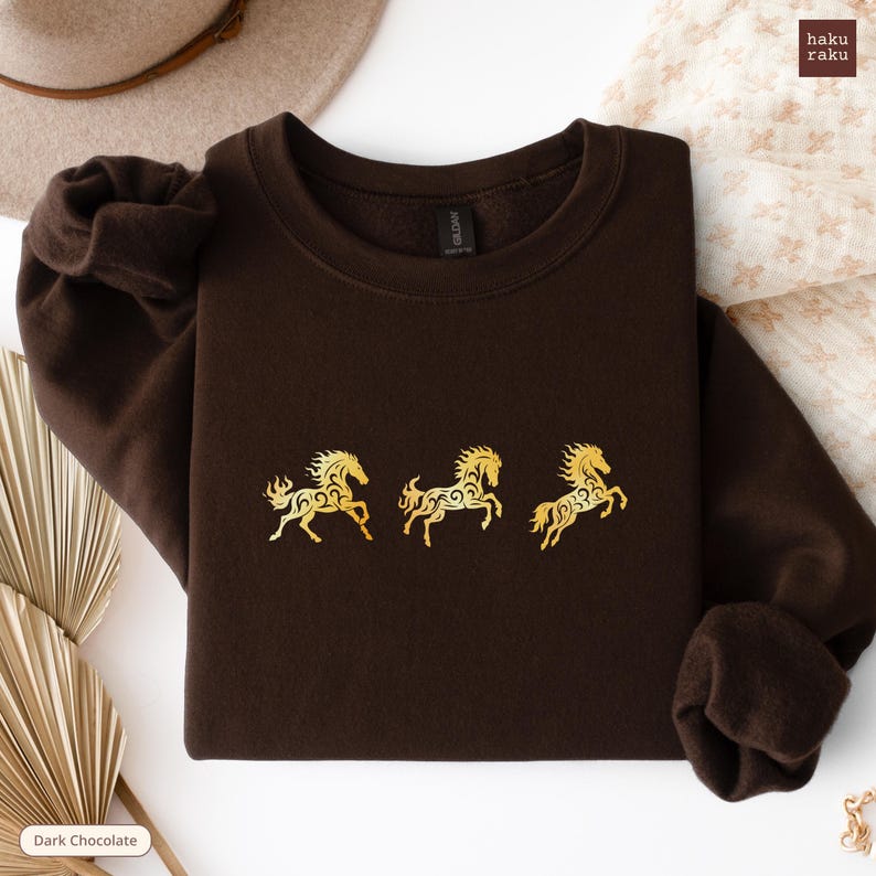 Op de afbeelding: Donkerchocoladekleurige sweatshirt met drie goudkleurige paardenontwerpen. De paarden zijn gestileerd met ingewikkelde patronen. De sweatshirt is opgevouwen, met de mouwen en halslijn zichtbaar. De merknaam "haku raku" staat rechtsboven.