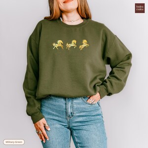 Op de afbeelding: Olijfgroene sweatshirt met drie gouden geborduurde paardenontwerpen. De crew neck sweatshirt wordt gecombineerd met een blauwe jeans. De merknaam "haku raku" staat rechtsboven. De tekst "Military Green" staat linksonder.