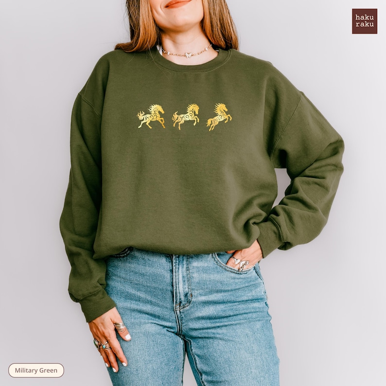 Op de afbeelding: Olijfgroene sweatshirt met drie gouden geborduurde paardenontwerpen. De crew neck sweatshirt wordt gecombineerd met een blauwe jeans. De merknaam "haku raku" staat rechtsboven. De tekst "Military Green" staat linksonder.