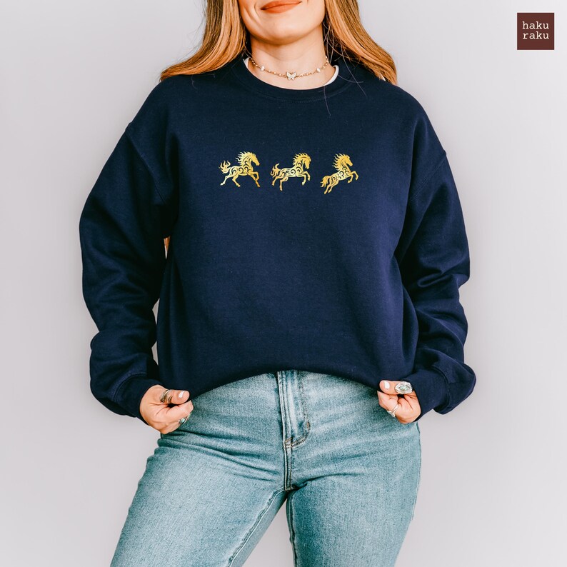 Op de afbeelding: Marineblauw sweatshirt met drie goudkleurige paardenmotieven op de borst. De crewneck trui is effen van kleur en wordt gecombineerd met een blauwe jeans. De merknaam "haku raku" staat rechtsboven.