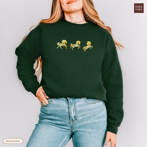 Op de afbeelding: Bostgroen sweatshirt met drie gouden paardenontwerpen. De trui heeft een ronde hals en lange mouwen. De tekst "haku raku" staat rechtsboven. De woorden "Forest Green" staan linksonder.
