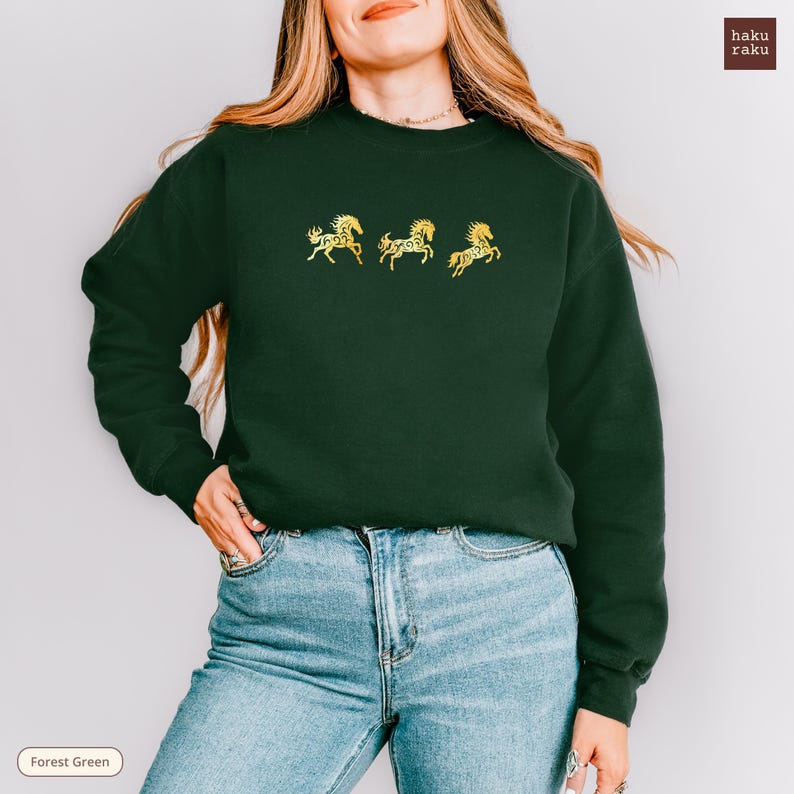 Op de afbeelding: Bostgroen sweatshirt met drie gouden paardenontwerpen. De trui heeft een ronde hals en lange mouwen. De tekst "haku raku" staat rechtsboven. De woorden "Forest Green" staan linksonder.