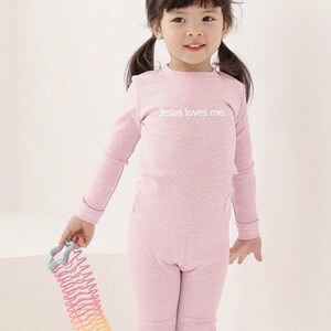 Puede incluir: Una ni&ntilde;a peque&ntilde;a con un conjunto rosa claro que consiste en una camisa de manga larga y pantalones a juego. La camisa tiene el texto "Jesus loves me". Un juguete de resorte arco&iacute;ris de colores se sostiene en la mano de la ni&ntilde;a.