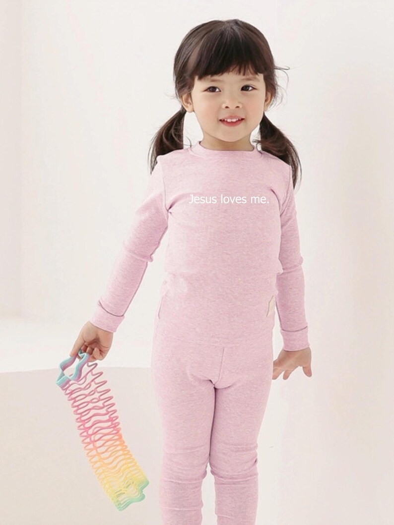 Puede incluir: Una ni&ntilde;a peque&ntilde;a con un conjunto rosa claro que consiste en una camisa de manga larga y pantalones a juego. La camisa tiene el texto "Jesus loves me". Un juguete de resorte arco&iacute;ris de colores se sostiene en la mano de la ni&ntilde;a.