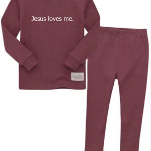 Puede incluir: Conjunto de pijama de dos piezas color granate. La parte superior de manga larga tiene el texto "Jesus loves me" en blanco. Los pantalones a juego tienen un dise&ntilde;o ajustado. Una peque&ntilde;a etiqueta est&aacute; cosida en la parte inferior de la parte superior.