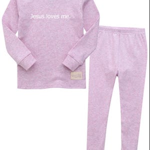 Puede incluir: Un conjunto de pijama lila claro con una parte superior de manga larga y pantalones a juego. La parte superior presenta el texto "Jesus loves me" en blanco. El conjunto est&aacute; hecho de un material suave y c&oacute;modo.