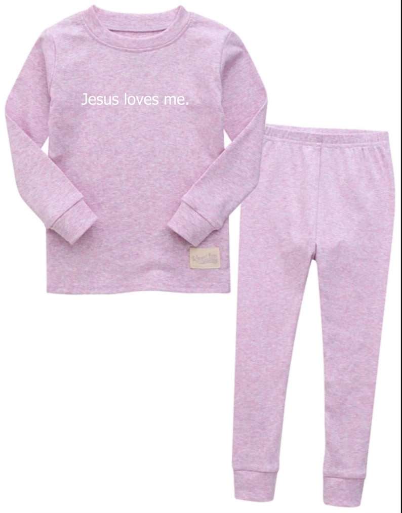 Puede incluir: Un conjunto de pijama lila claro con una parte superior de manga larga y pantalones a juego. La parte superior presenta el texto "Jesus loves me" en blanco. El conjunto est&aacute; hecho de un material suave y c&oacute;modo.