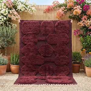 May include: A deep burgundy rectangular rug featuring a shaggy, high-pile texture. The surface is decorated with a repeating geometric pattern of interconnected hexagonal and rounded shapes, creating a raised, tactile design across the fabric.