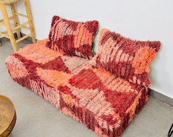 May include: A vibrant, hand-woven Moroccan-style floor sofa in shades of red, orange, and pink. The sofa features two plush cushions and a low-profile design. The textured fabric adds visual interest and comfort.