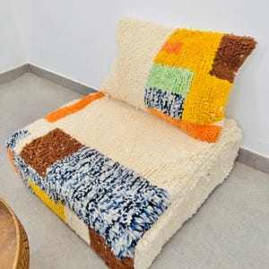 Puede incluir: Un conjunto de cojín de suelo y almohada con un diseño de patchwork. El cojín y la almohada presentan una base crema con secciones de naranja, marrón, amarillo, azul y verde. La tela texturizada añade atractivo visual al conjunto.