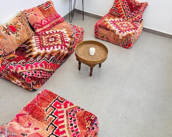 May include: A living room set featuring a two-seat sofa, a single-seat chair, and a footstool, all with vibrant red, pink, and orange geometric patterns. A small, round wooden side table and a decorative tray complete the scene.