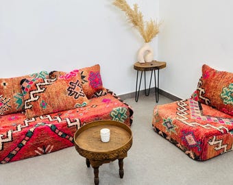 May include: A living room set featuring a sofa and armchair with vibrant, patterned upholstery in shades of red, orange, and pink. A small, round wooden table with carved details and a white candle sits in front of the sofa. A side table with a vase and dried flowers is also present.