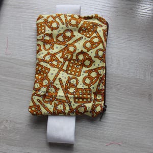 May include: A rectangular fabric pouch featuring a repeating pattern of brown pretzels on a light yellow background with white salt speckles. The pouch includes a dark brown zip closure and a wide white elastic band attached to the top and bottom.
