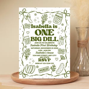 May include: A white invitation card with a green border and text. The text reads "Isabella is ONE BIG DILL" with details for a first birthday party, including the date, time, and location. Decorative pickle illustrations surround the text.