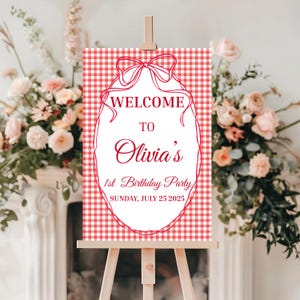 May include: A red and white gingham patterned welcome sign on a wooden easel. The sign has a decorative red border and a bow at the top. The text reads "WELCOME TO Olivia's 1st Birthday Party Sunday, July 25 2025."