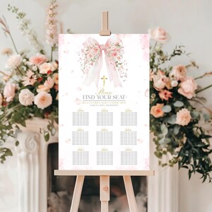 Op de afbeelding: Een wit tafelplanbord op een houten ezel met een grote roze aquarel strik, bloemaccenten en een gouden kruis. De tekst luidt: Please FIND YOUR SEAT, BELLA'S FIRST HOLY COMMUNION, MARCH 2 20XX, MELBOURNE, en bevat een lijst met tafels.