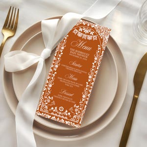 May include: An orange menu card with white floral and banner designs, featuring the text "Menu" and details for "The Rodriguez Baby Shower." The menu lists a starter, main course, and dessert. A white ribbon is tied around the card, placed on a plate with a gold fork and knife.