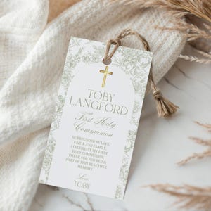 Puede incluir: Una etiqueta blanca con una cruz dorada y un diseño floral, atada con hilo. La etiqueta dice "TOBY LANGFORD Primera Santa Comunión" con un mensaje debajo. La etiqueta está sobre una tela blanca.
