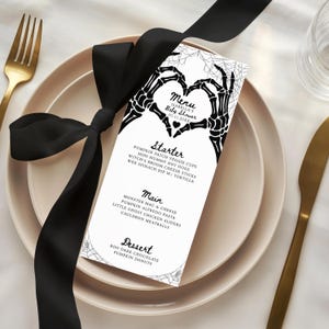 May include: A white rectangular menu card featuring black skeleton hands forming a heart shape at the top, with spider web illustrations in the corners. The text reads Menu, Isabella's Baby Shower, 01.11.20XX, Starter, Main, and Dessert with food items.