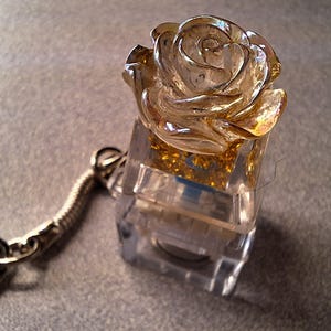 May include: A clear resin keychain featuring a golden rose encased in a clear, square block. The rose has detailed petals and sits atop a base filled with gold glitter. A silver-colored metal chain and clasp complete the accessory.