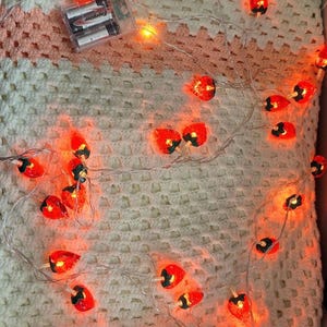 May include: A string of decorative lights shaped like strawberries, with red and green details, illuminated against a white and peach crocheted blanket. The lights are powered by batteries.