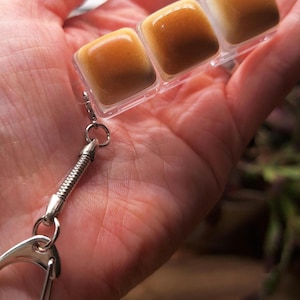 May include: A keychain featuring three miniature, square-shaped, toasted marshmallow-like charms encased in a clear plastic frame. The charms have a gradient of golden-brown to white coloring. A silver-toned metal chain and clasp are attached.