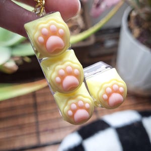 May include: A yellow and pink paw print charm on a gold chain. The charm features three paw prints in a row, with pink pads and yellow toes. The charm is attached to a gold ball chain.