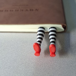 May include: A brown book with a whimsical bookmark featuring black and white striped legs and red shoes. The bookmark is inserted between the pages, adding a playful touch to reading.