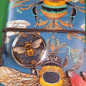 May include: A blue journal with a bee illustration and a round, silver-rimmed compact mirror featuring a bee design. The journal is secured with a brown cord. The bee design is yellow, black, and orange.