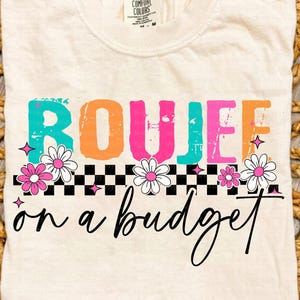May include: Cream-colored t-shirt with the words "BOUJEE on a budget" in colorful, playful lettering. The design includes a checkerboard pattern, flowers, and star accents, creating a fun and stylish look.