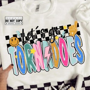 Può includere: Felpa bianca con la scritta "Let's go TORNADOES" in lettere colorate e bordate. Il design include una faccina sorridente, fulmini e una mano con il numero uno. Sono visibili anche le parole "designwinco" e "digital download".