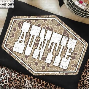 May include: Black t-shirt with a distressed, camouflage-patterned graphic that reads "AUNTIE" in bold, white letters. The shirt is folded and rests on a leopard print fabric. The image also includes disco balls.